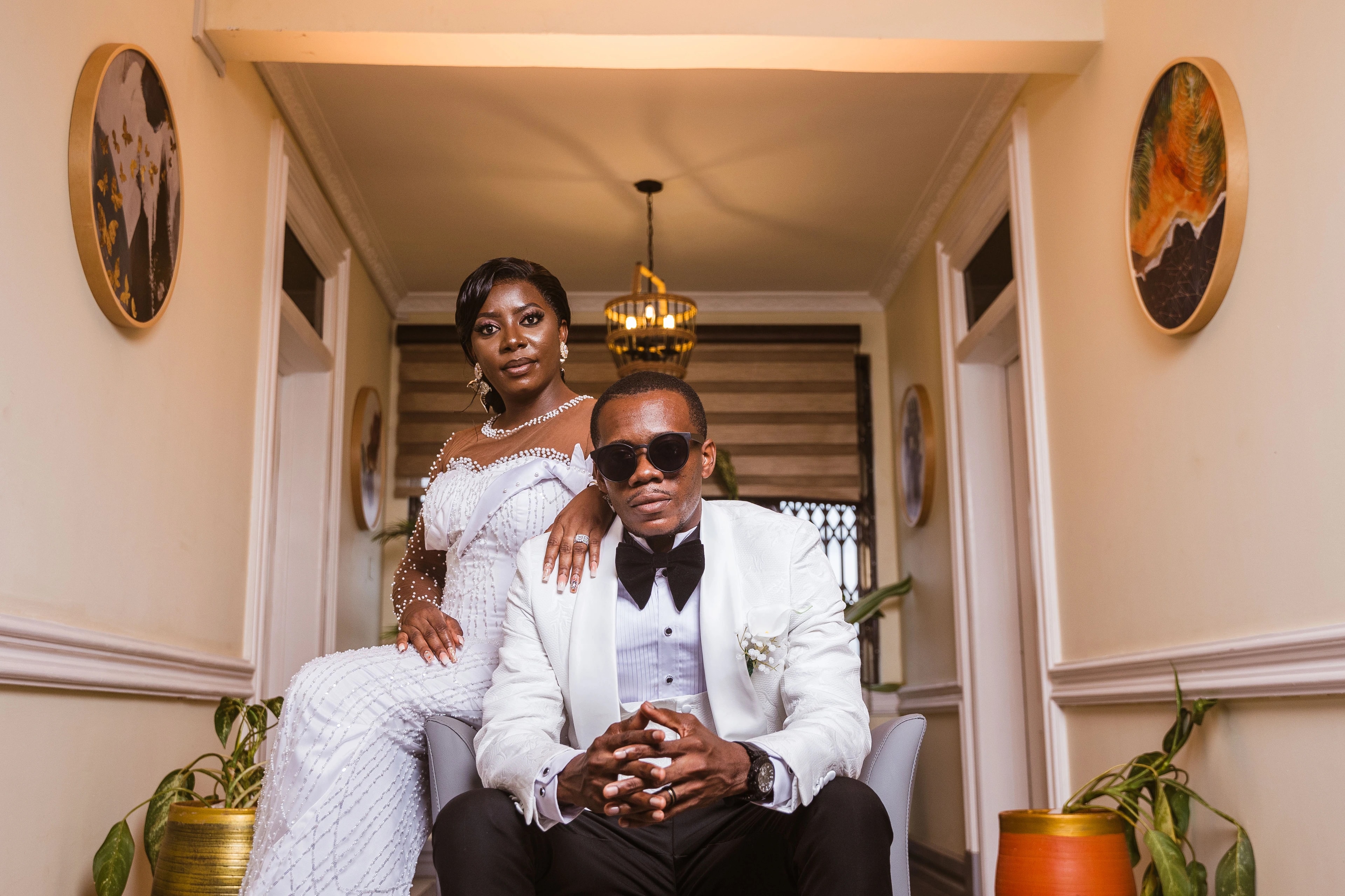 A bride stands behind her seated groom in a warmly lit hallway